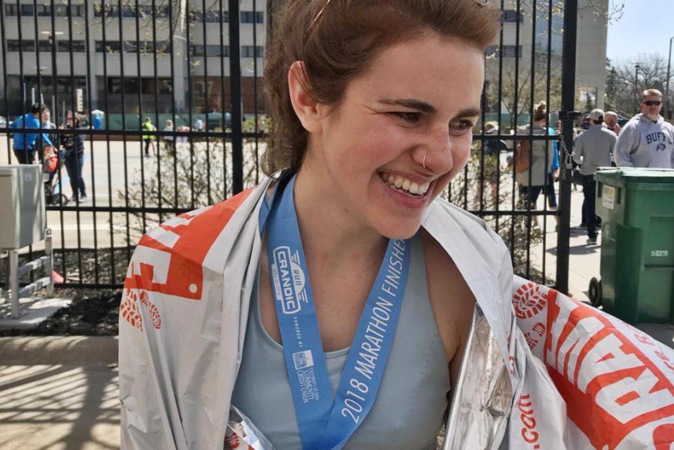 Lucia smiles gratefully wearing a 2018 Marathon Finisher medal around her neck and a marathon flag wrapped around her shoulders