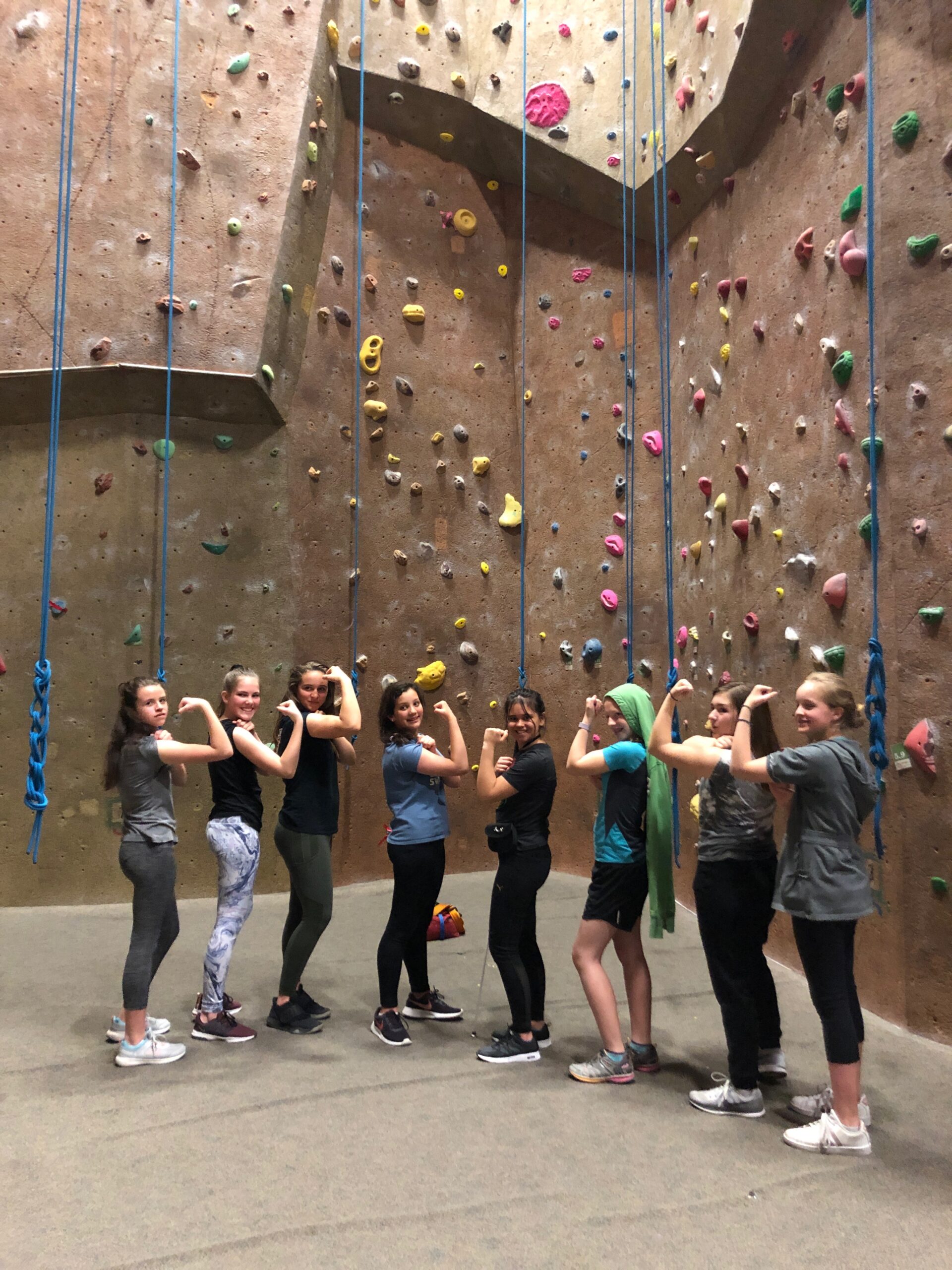 A group of girls stands in a line in front of a rock climbing gym's rock wall flexing their arms