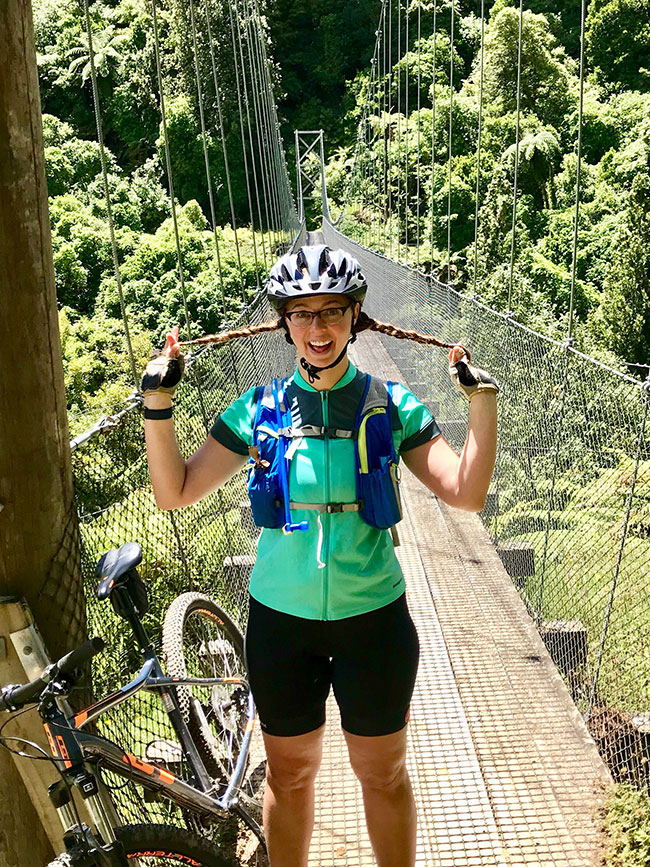 Colette Roy stands on a bridge with a mountain bike leaned against the railing; she's holding her braids out to the side and smiling big
