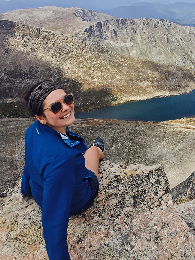 Cairn Project Ambassador Olivia Grev looks back and smiles wearing sunglasses sitting on a rocky mountain peak