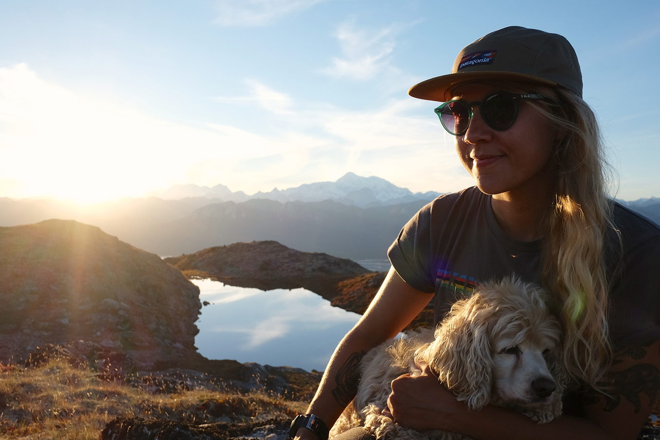 Emily sits on a mountaintop with her dog in her lap as the sun sets behind them