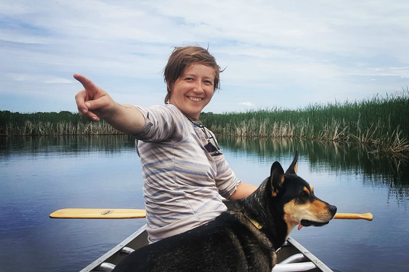 Siri sits in a canoe with a dog and points behind her smiling