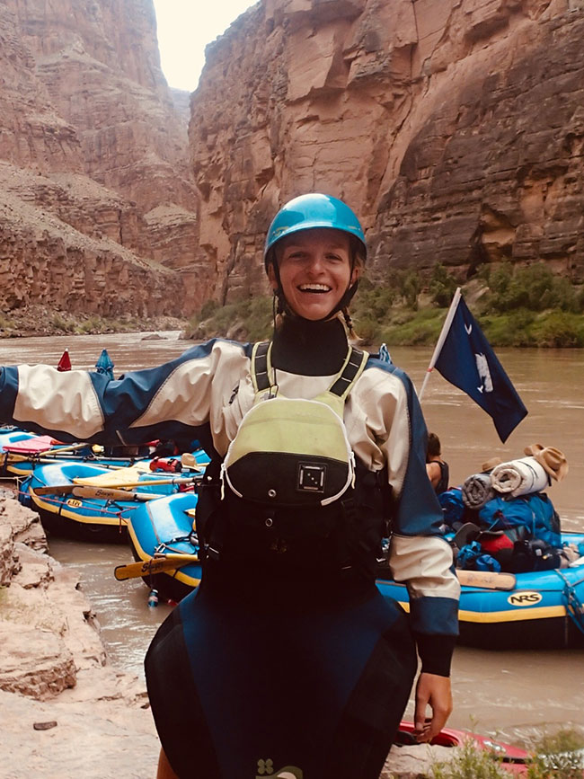 Laura Wildenborg smiling while wearing full raft gear with rafts and tall orange desert walls behind her
