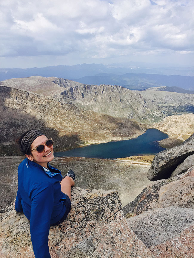 Olivia Grev smiles at the camera from a mountain peak with rocky mountain ridges and an alpine lake in front of her