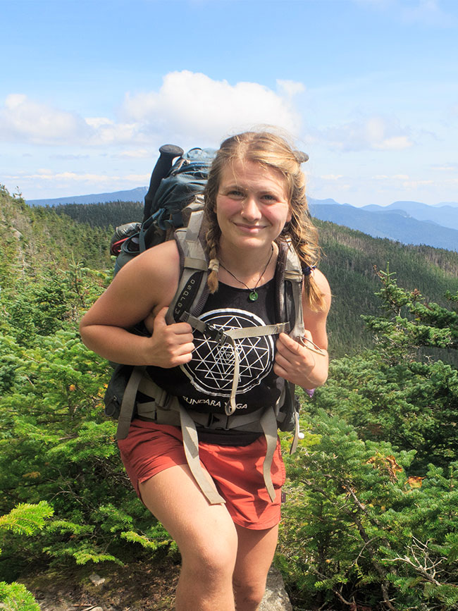 Cairn Project Ambassador Emma Corwin smiles wearing a backpacking pack with mountain ridges and pine trees behind her
