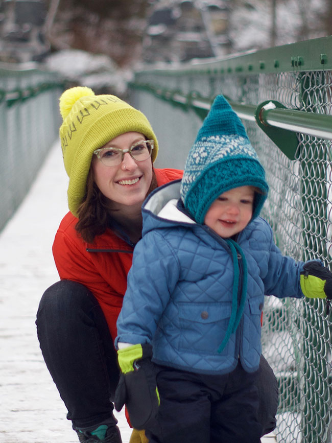 Cairn Project Ambassador Emma Schultz smiles in a yellow beanie on a pedestrian bridge while crouched behind her child who's bundled up for the winter weather