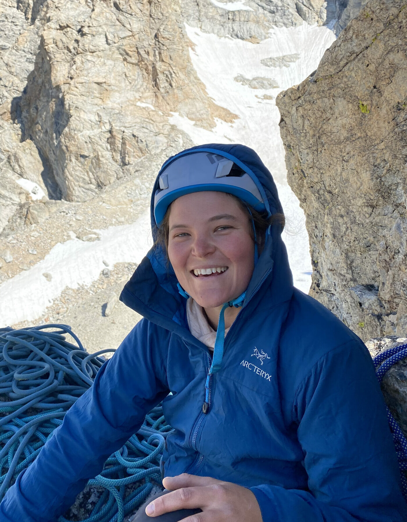 Cairn Project Ambassador Leah Genth smiles on a multipitch climb with rope coiled beside her and a snowy mountainside behind her