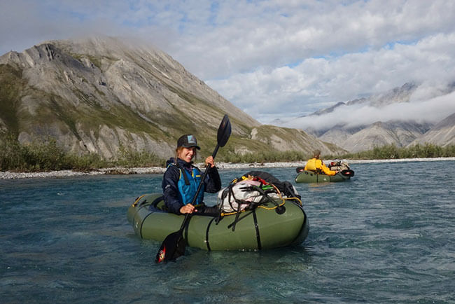 Molly paddles a packraft on a mountain lake with foggy clouds over the rocky mountains in the background