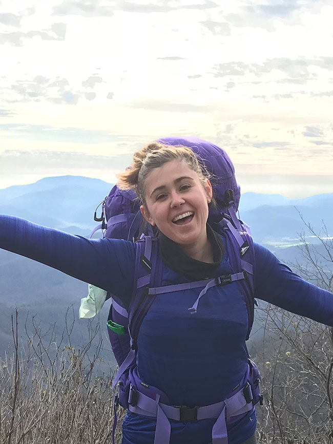 Cairn Project Ambassador Haleigh Ehmsen wearing a purple shirt and a purple backpack smiles with her arms held out