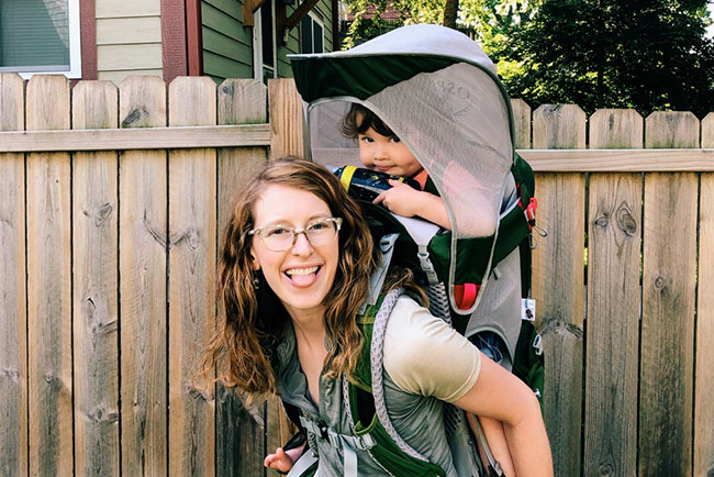 Emma smiles and sticks her tongue out with her child in a backpack on her back