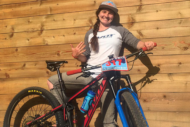 Kate smiles while holding her mountain bike against a wooden wall