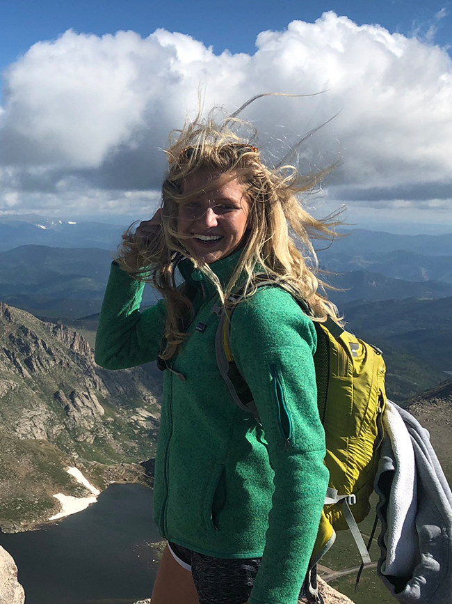 Cairn Project Ambassador Lydia Terhaar smiles on a mountaintop with her hair blowing in the wind