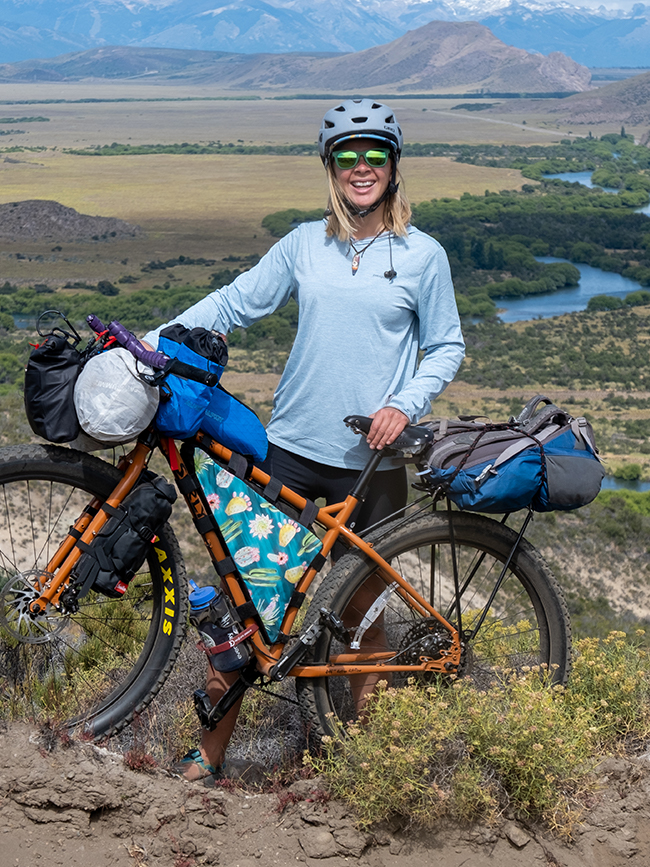 Cairn Project Ambassador Becca Foss smiles holding her bike packed full of bike touring supplies with mountains in the background
