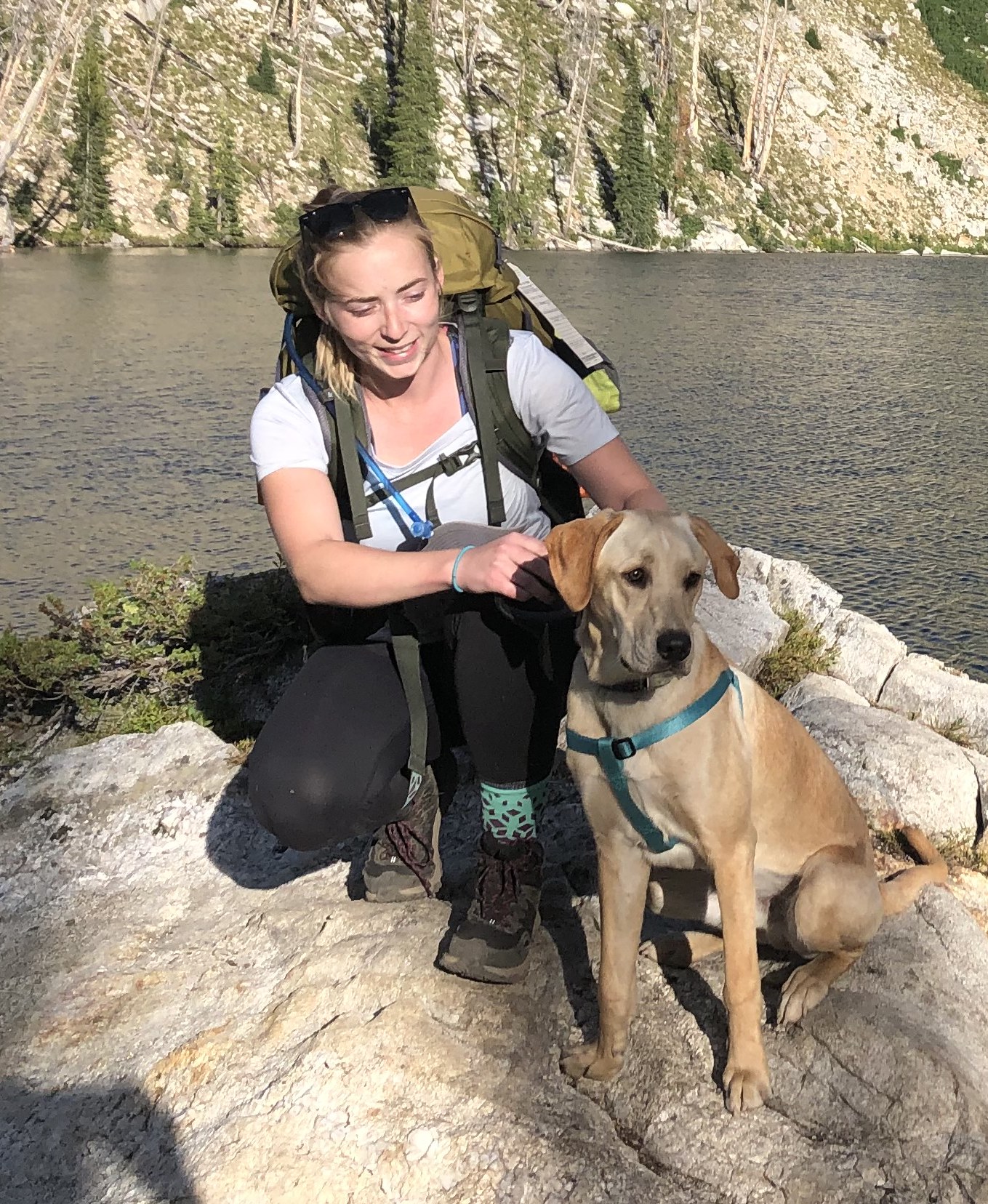 Cairn Project Ambassador Shelby McGibney wears a backpack next to an alpine lake with her yellow lab