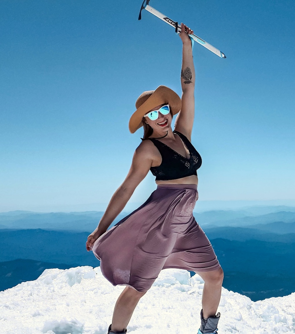 Cairn Project Ambassador Angie Marie holds an ice axe in the air on a snowy mountaintop while wearing a pink skirt, black top, sunglasses, and sun hat