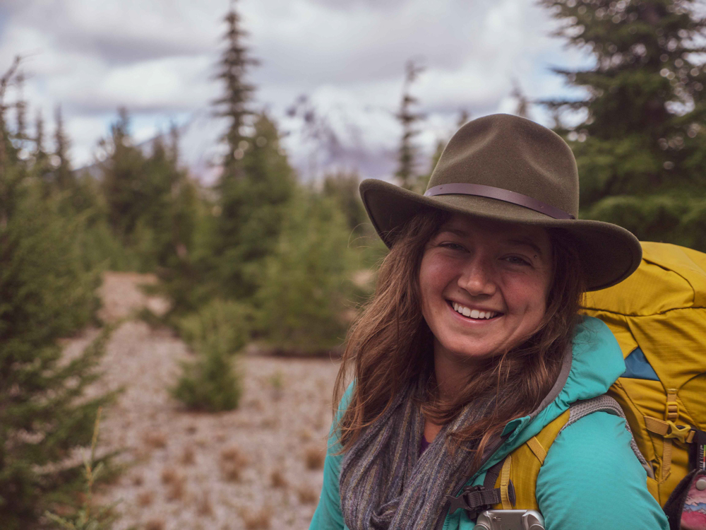 Roxy smiles wearing a brown hat and. yellow backpacking pack with mountains and pine trees in the background