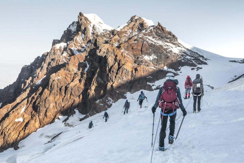 A group of AWExpeditions guests make their way across a snow-covered hill with ice axes and crampons with a big mountain peak in front of them