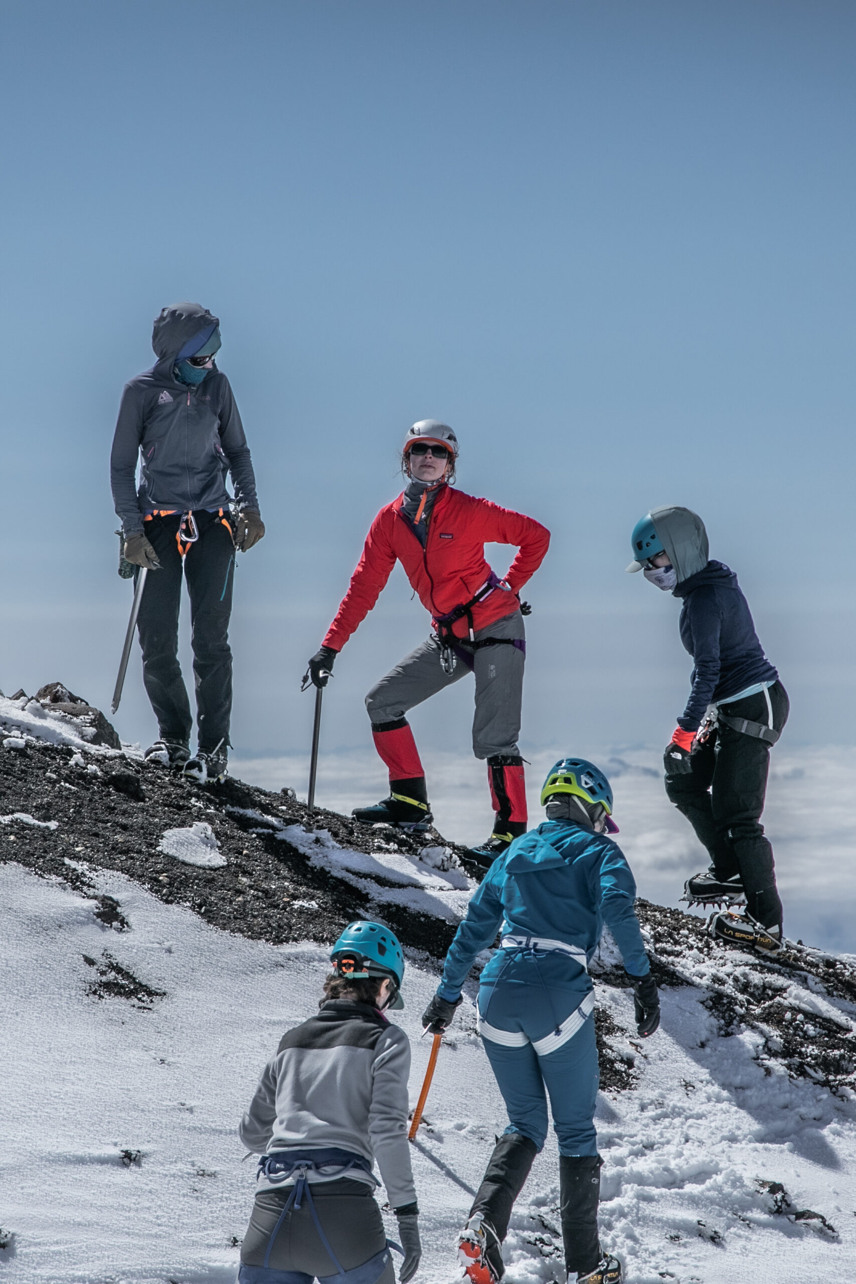 Five women mountaineers walk up a snow-covered mountain ridge holding ice axes and wearing harnesses; a sea of clouds is behind and below them in the background