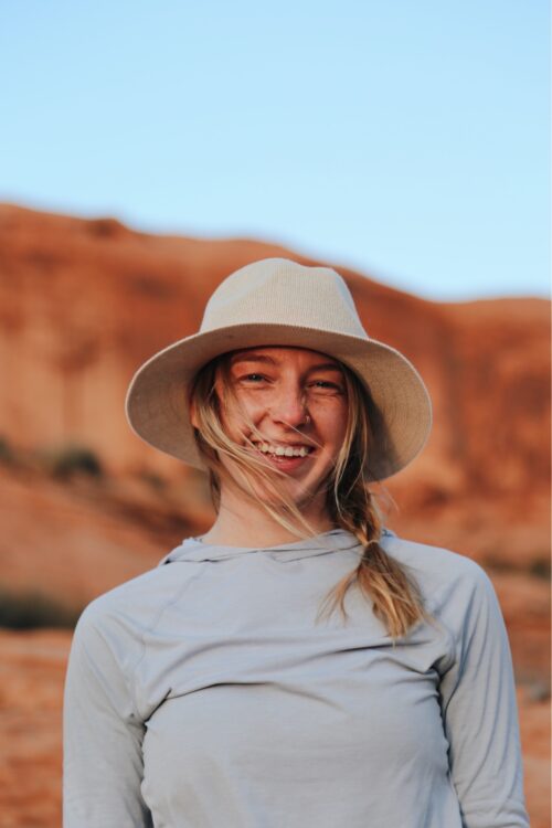 Cairn Project Ambassador Abbie Cheney smiles wearing a sun hat with her blonde hair in her face and red desert rock behind her
