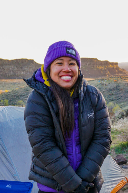 Cairn Project Ambassador Sydney Pham smiles wearing two puffy coats in front of a tent and red desert rocks