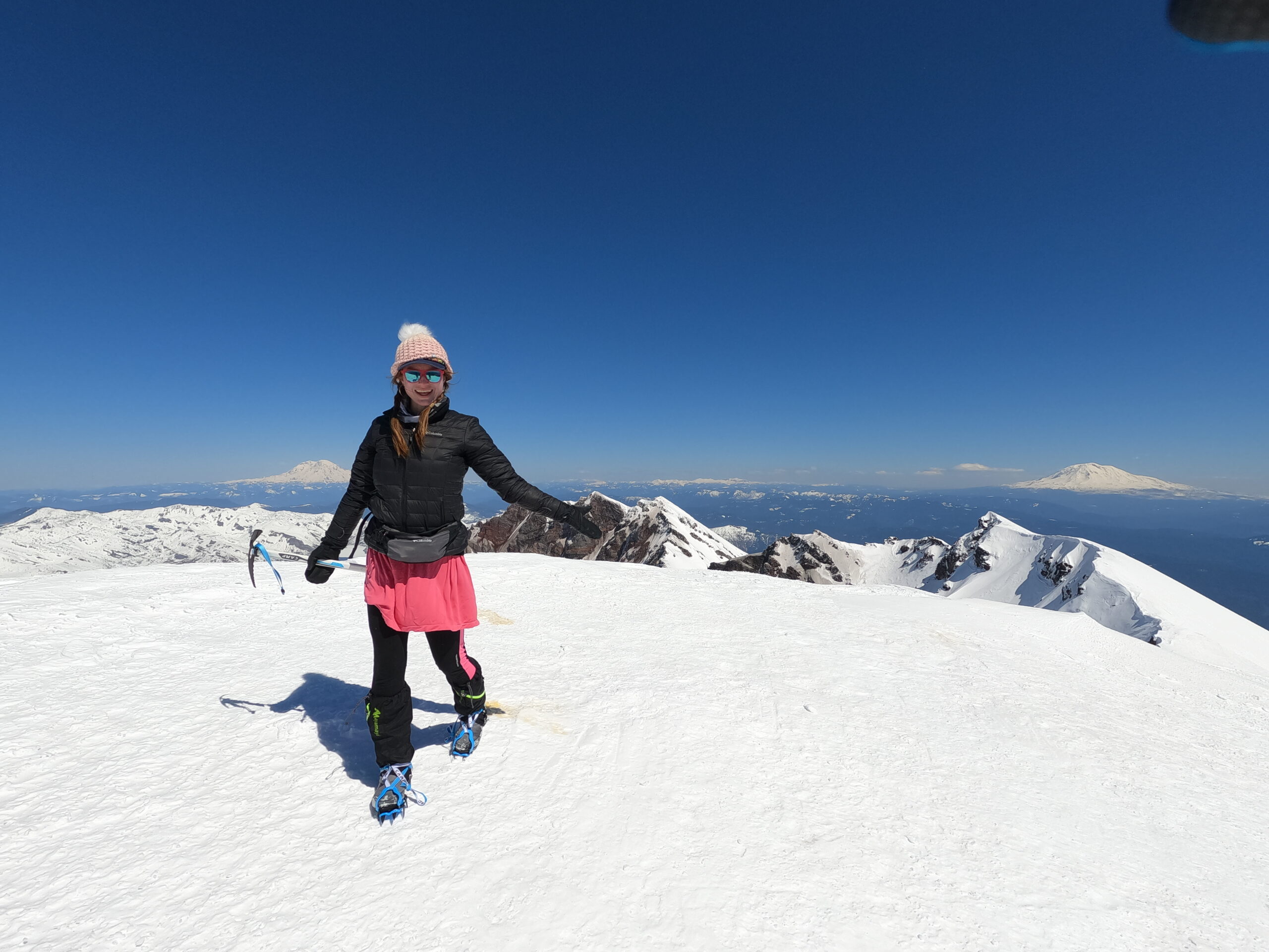 Cairn Project Ambassador Angie Marie holds an ice axe on a snowy mountaintop while wearing a pink skirt, black puffy jacket, crampons, and beanie