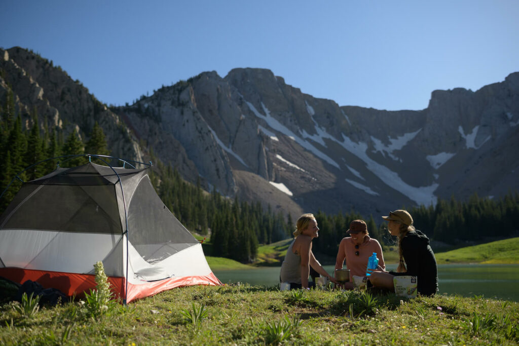 three women sit by a tent in front of a steep mountain range