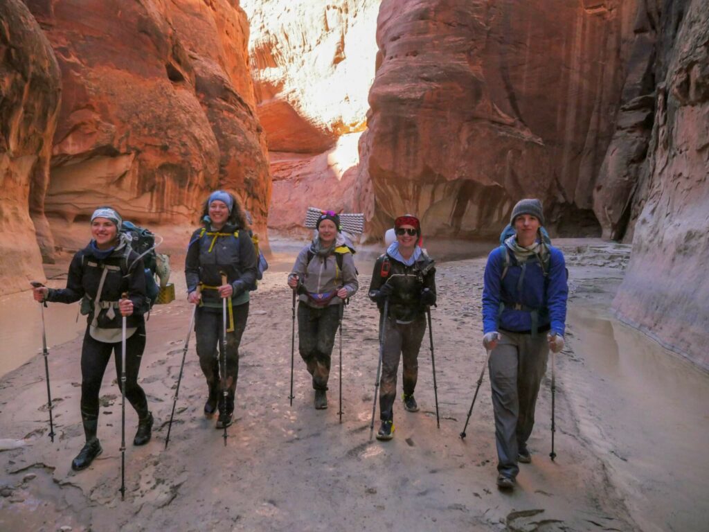 five women walk toward the camera in a canyon wearing backpacking gear