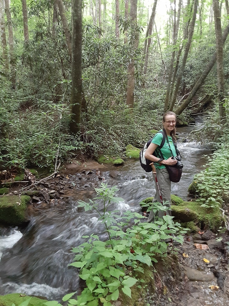 Alessandra Trompeo stands next to a river with hiking clothes on and trees towering in the background