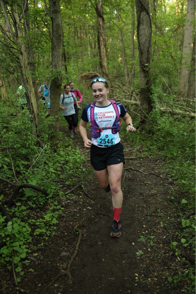 Dana Ringler wearing a white shirt and running vest running toward the camera and smiling. there are woods and other runners behind her