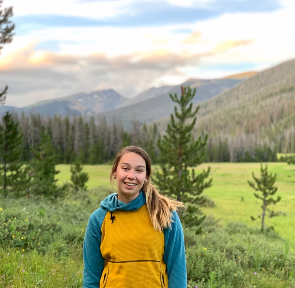 cairn project ambassador kelly gazarik smiles at the camera wearing a melezana sweatshirt with green mountains and blue sky behind her