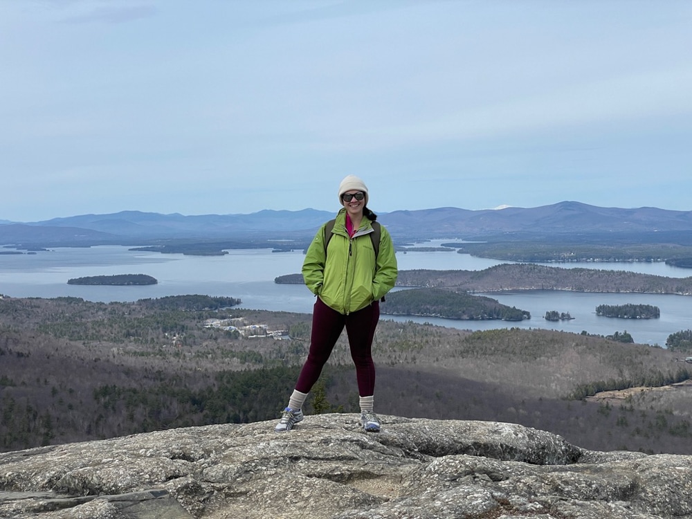 Suleyka Garcia Rivera stands at the top of a mountain smiling at the camera, with lakes and islands behind her