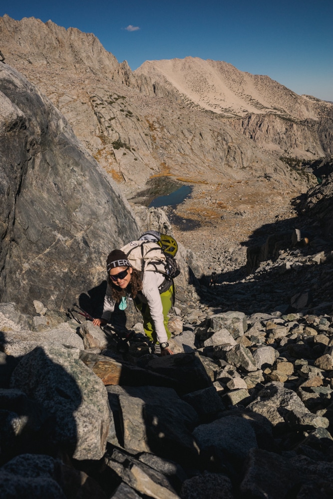 Viv Serrano climbs a boulder field toward the camera weating sunglasses and a aht