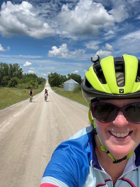Kathy Murph smiles to the camera in the foreground in a helmet and bike jersey. In the background there are two other bikers on a long dirt road
