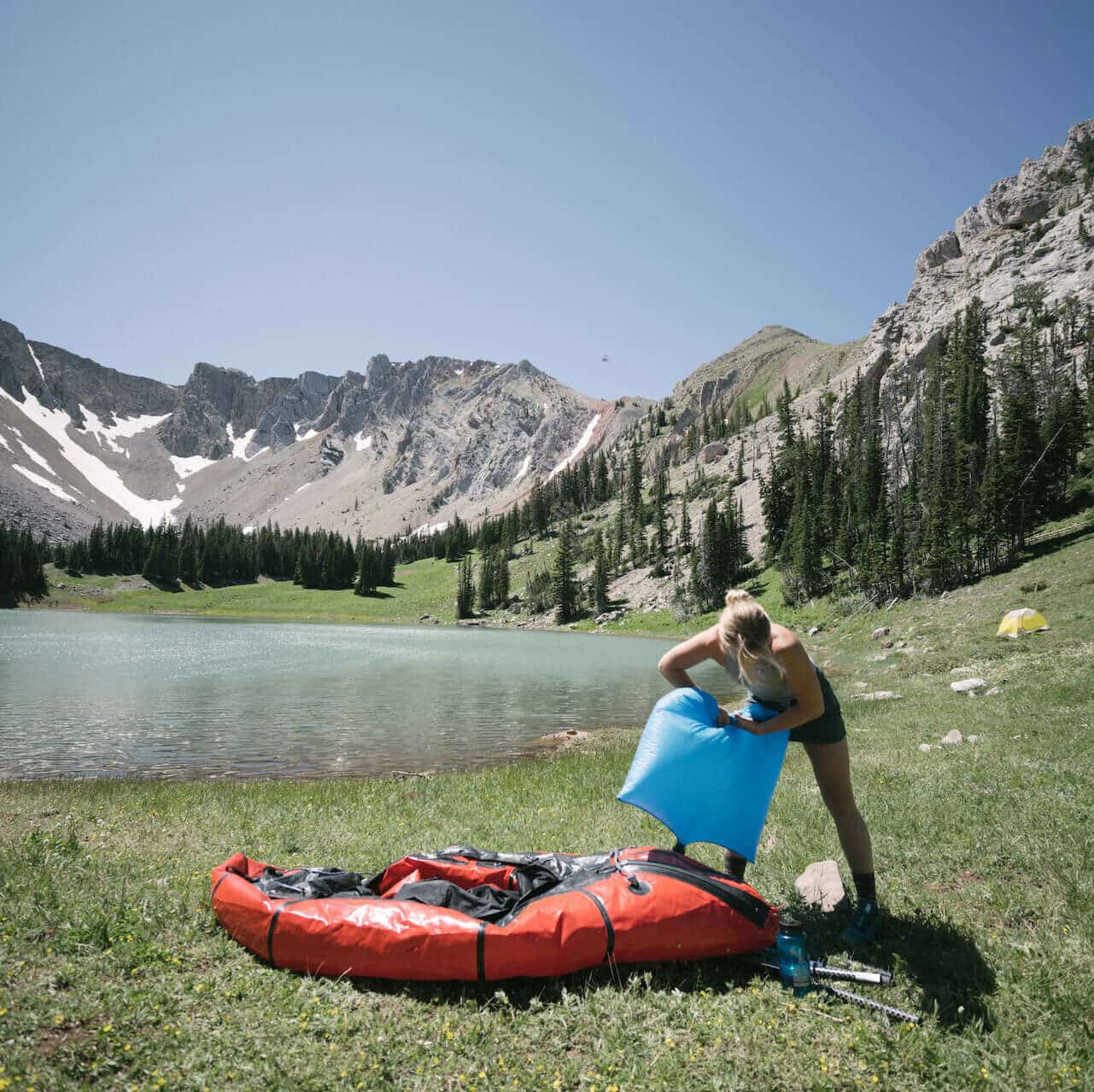 A Cairn Project ambassador pumps up a red inflatable kayak with an alpine lake and mountain peak behind her