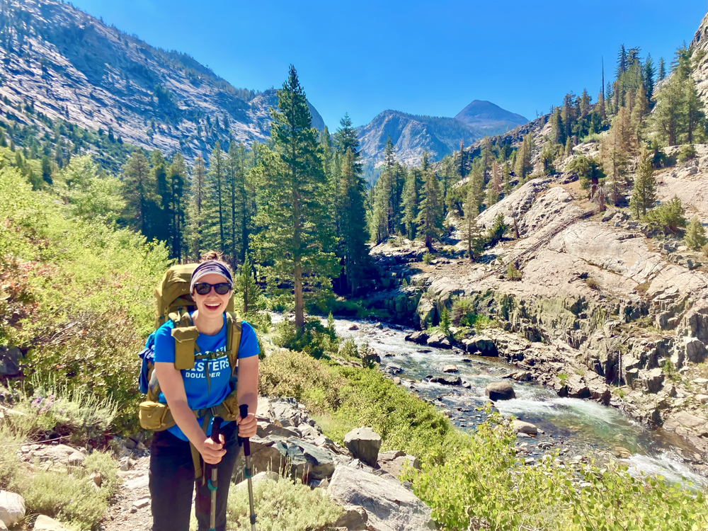 Kelly smiles towards the camera with a stream and moutains behind her. She is carrying a backpack and wearing hiking clothes.